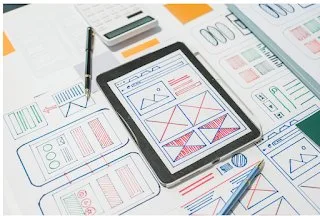 Hand-drawn screen layout sketches on paper around a tablet showing a matching wireframe