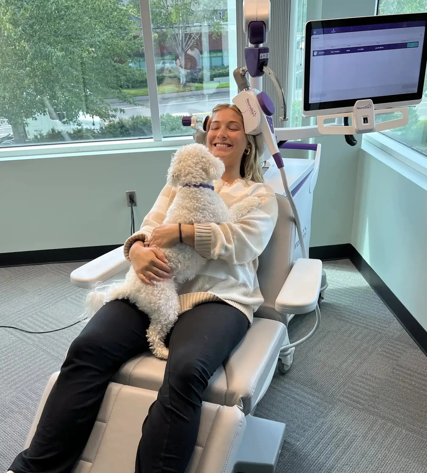 Patient holding a small white dog while seated under a NeuroStar TMS coil in a sunlit clinic room