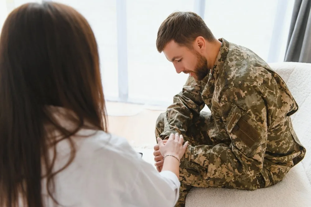 Clinician resting a hand on the clasped hands of a service member in camouflage uniform